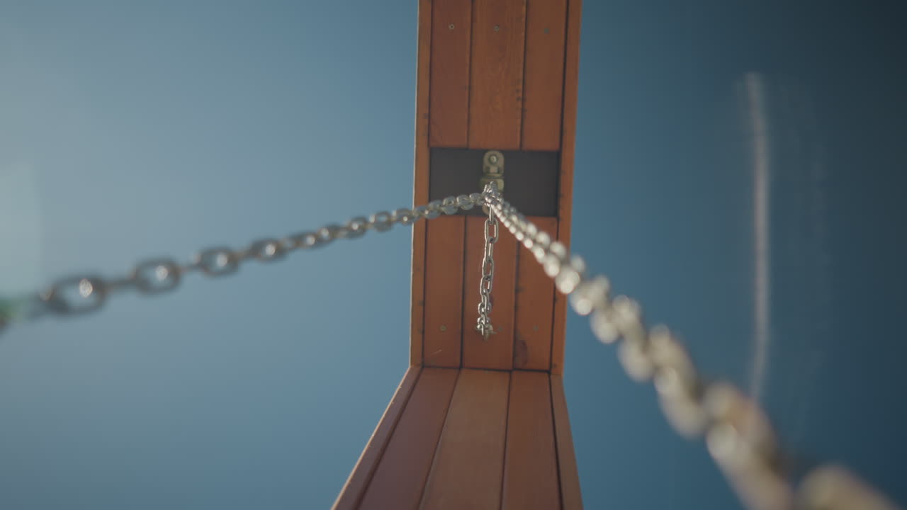 나무 프레임에 부착된 금속 사슬의 클로즈업, 밝은 파란 하늘을 배경으로 낮은 각도에서 포착, 산업의 힘과 자연 재료가 야외 풍경과 여있는 것을 보여줍니다.