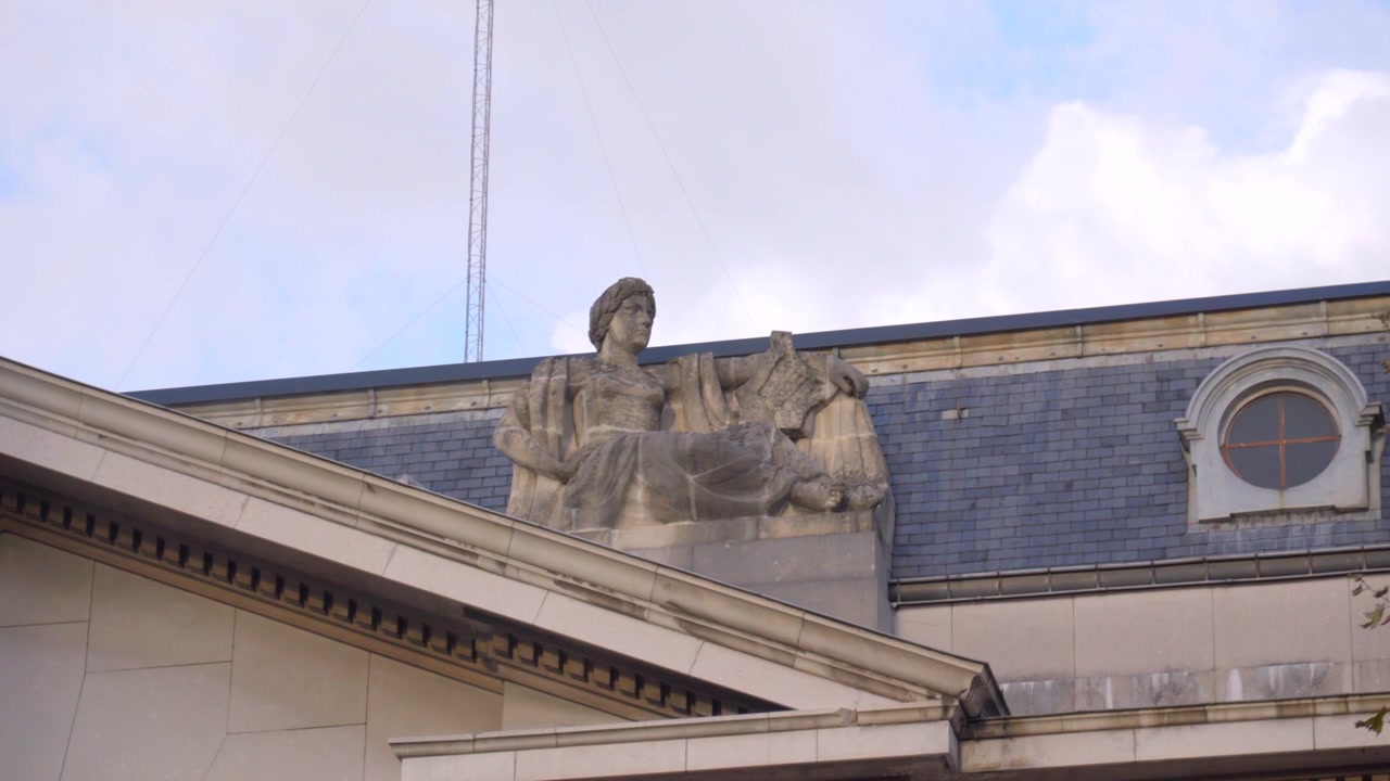 Una vista dal basso di una statua in pietra degli anni '30 di Claire Colinet (1885-1972) su un edificio amministrativo. I dettagli della statua risaltano contro il cielo coperto, filmata con una lunga lunghezza focale.