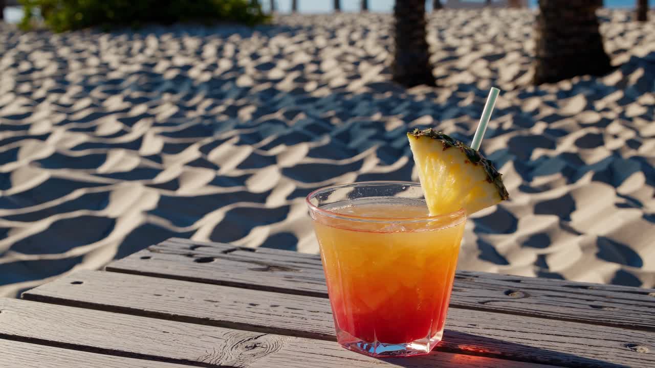 Six identical tequila sunrise cocktails, garnished with a pineapple wedge and a straw, sit on a weathered wooden table against a backdrop of pristine white sand and palm trees