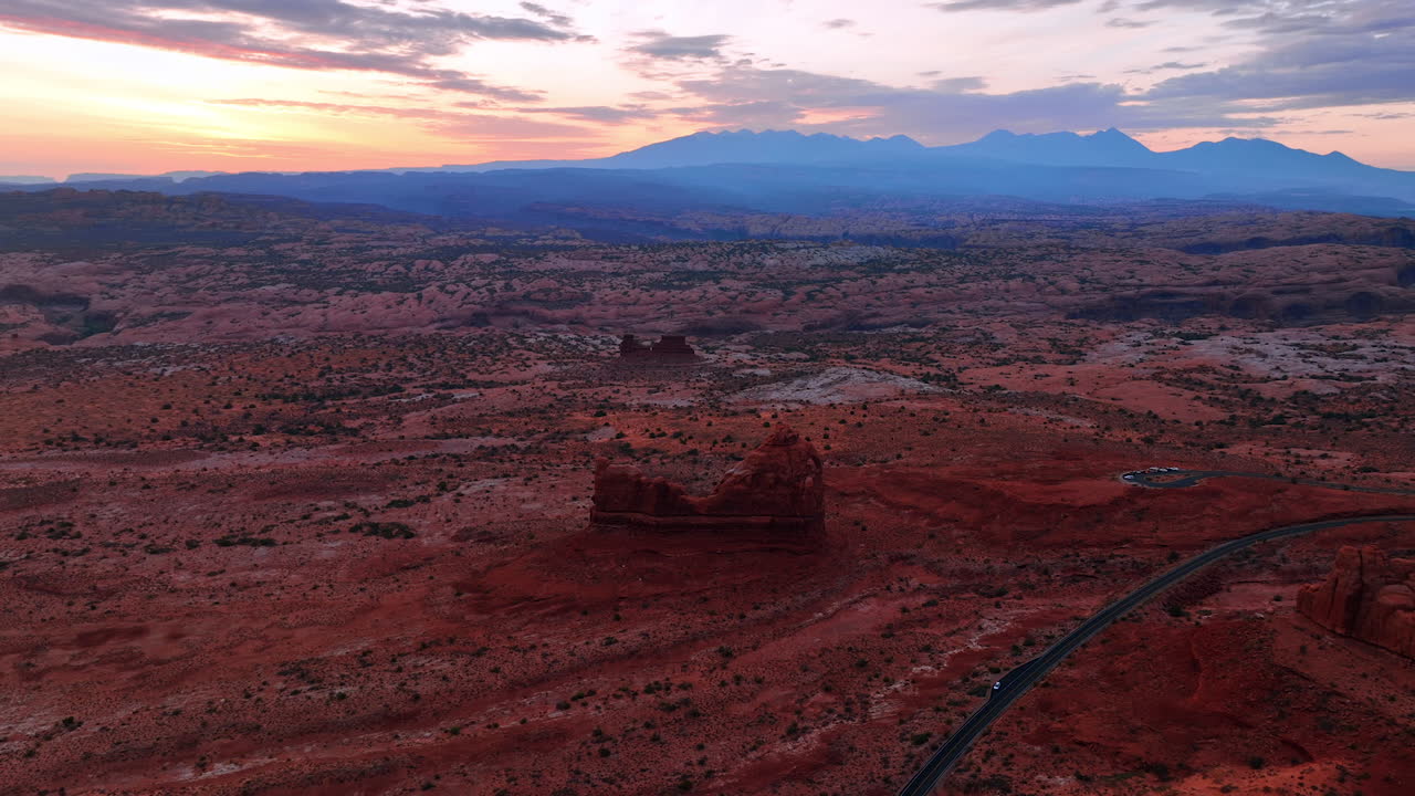 Arid deserted landscape with some shrubs growing in the scenery. White car stands parked at the road crossing the view. Mountains range silhouette at backdrop