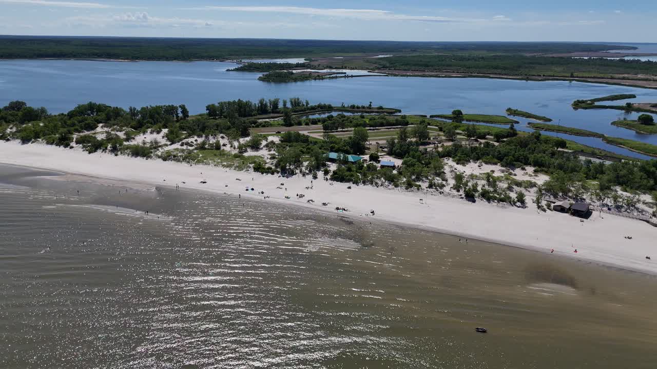 Kaukaa otettu ilmakuva Winnipeg-järven yltä itään kohti Grand Beachia Manitobassa aurinkoisena kesäpäivänä. Kamera pyörii oikealle paljastaen hiekkarannan, uimarit, laguunin ja havumetsän