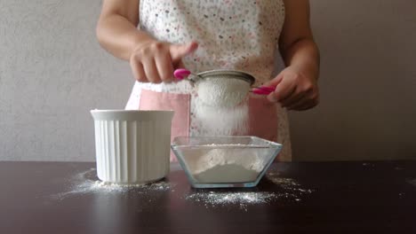 Mujer-Latina-Con-Un-Delantal-Preparando-La-Cocina-Horneando-Un-Pastel-Tamizando-La-Harina-Con-Un-Colador-De-Metal-Y-Su-Mano