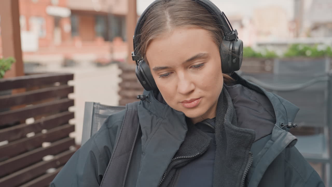 Donna caucasica che legge all'aperto indossando cuffie, sorseggiando un caffè sulla terrazza di un bar in città, sfogliando le pagine di un libro tascabile mentre indossa una giacca scura, espressione pensierosa, primi piani con illuminazione soffusa