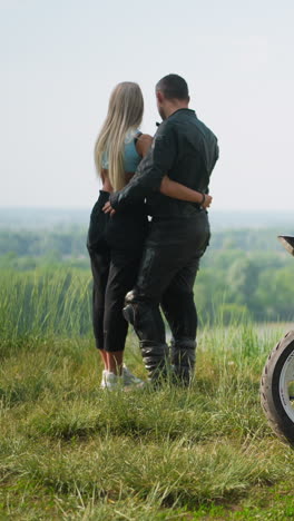 motorcyclist with slim girlfriend stands near contemporary bike on steep hill against picturesque lake on spring day backside view slow motion
