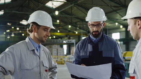 three engineers wearing helmets and holding blueprint while talking in a factory