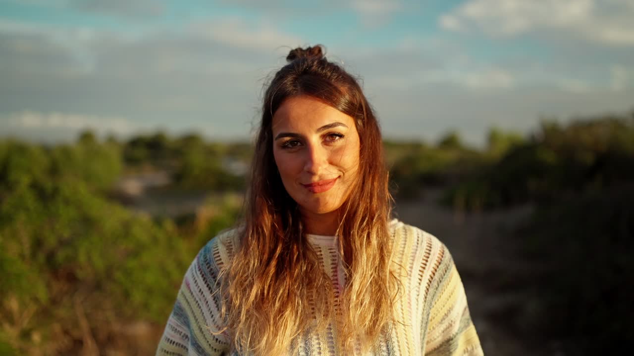 Movimiento de cámara lenta hacia una modelo de pie y sonriendo en la naturaleza al atardecer.