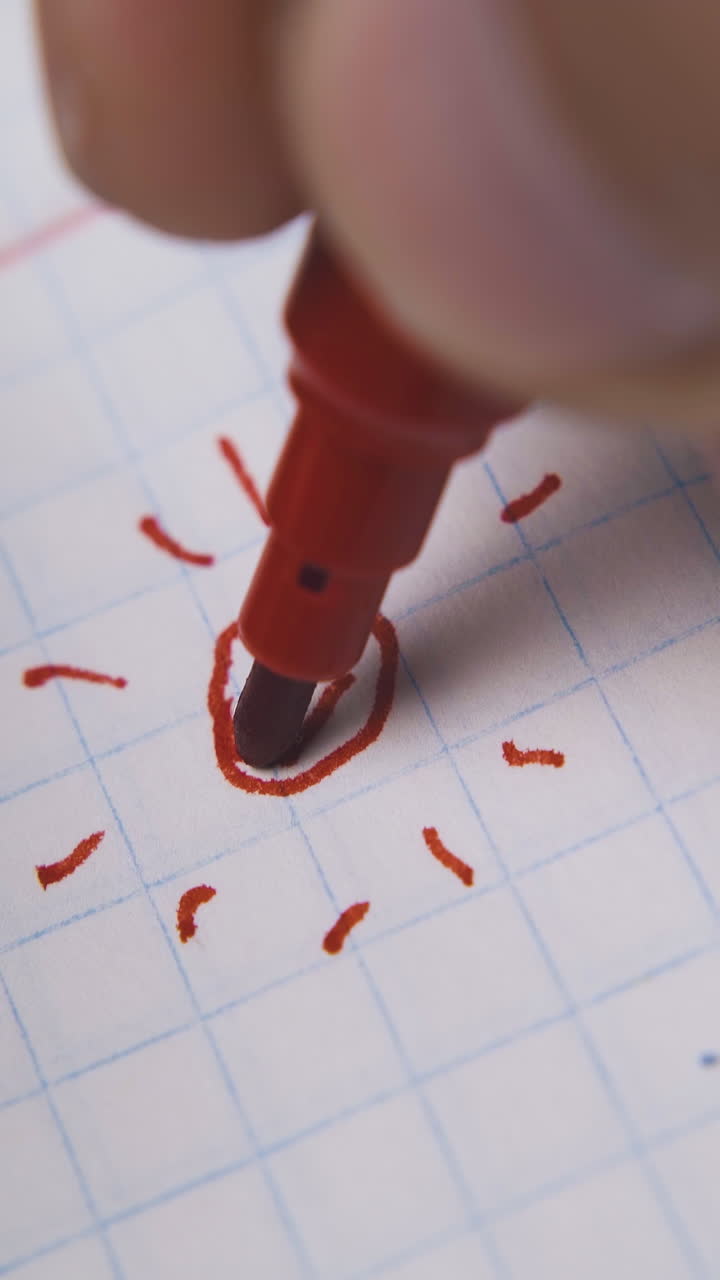 young man draws smiling sun using red marker on blank checkered paper sheet extreme close view