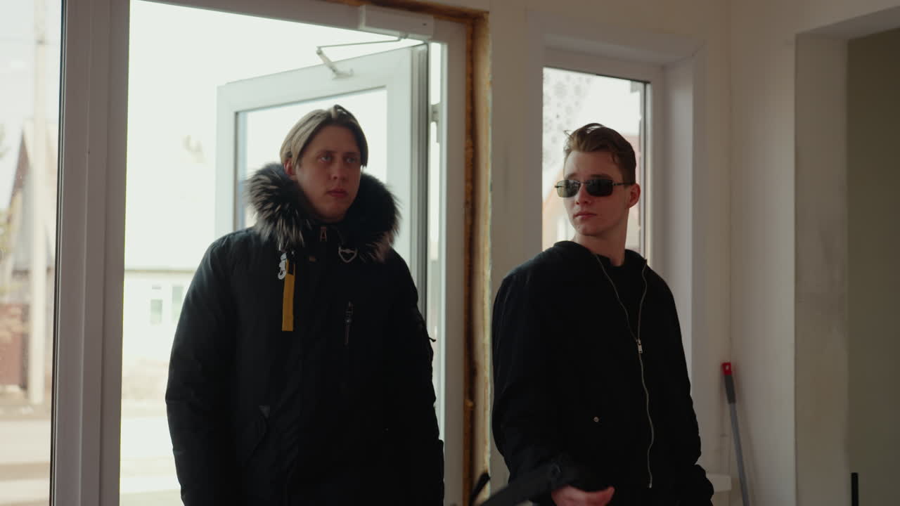 Men in sunglasses with black jacket and backpack enters through glass door while another man in winter coat follows behind, exterior suburban houses visible, moment of casual arrival captured indoors