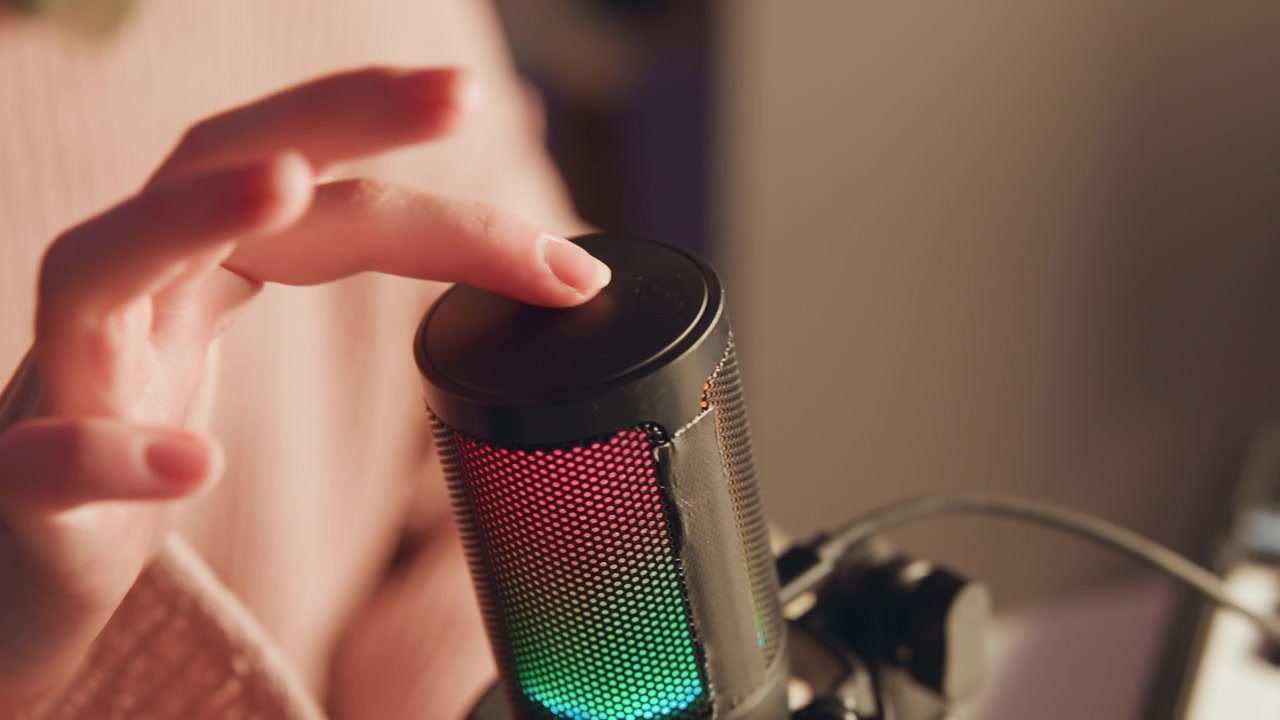 Warm studio scene showing illuminated condenser microphone with multicolor LED grille on shock mount, angled toward blurred speaker in pink sweater, with soft bokeh, desktop setup, and ambient
