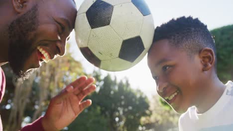 African-american-dad-and-son-balancing-the-football-together-with-their-heads-in-the-garden