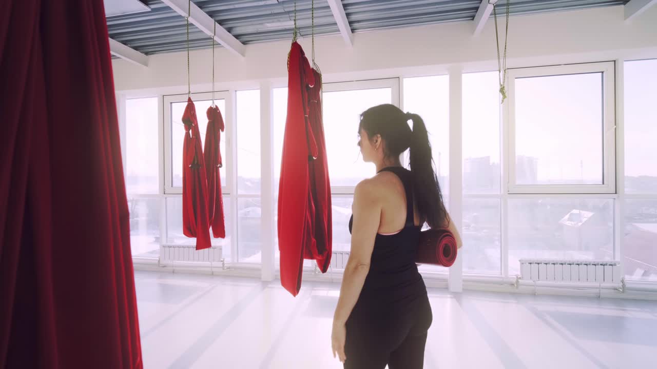 A poised woman stands in a bright, airy studio, preparing for a rejuvenating yoga session with a mat in hand and vibrant red garments hanging around her