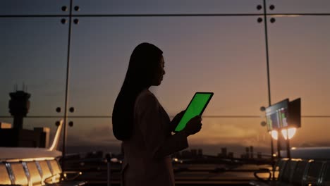 vista trasera de una mujer de negocios asiática con una maleta rodante en la sala de embarque del aeropuerto, usando una tableta de pantalla verde mientras espera el vuelo, el avión despega fuera de la ventana