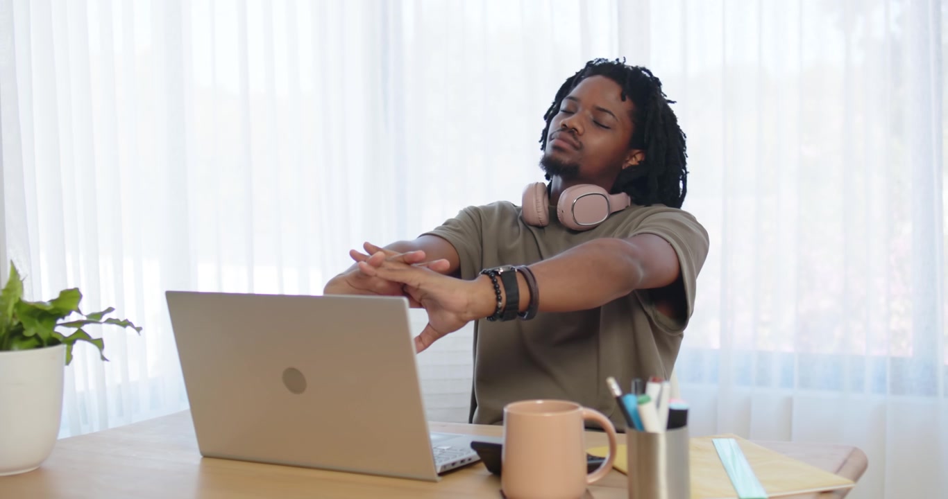 Adult man taking seated stretch break after laptop work at desk, leaning back and rotating neck