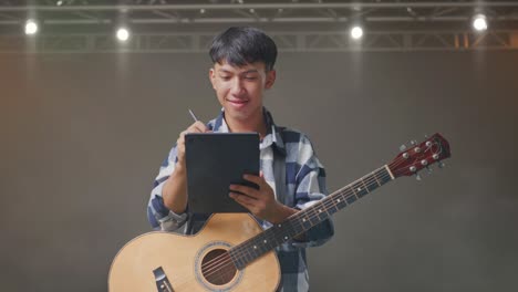 asian teenager with guitar taking note on a tablet while standing on stage