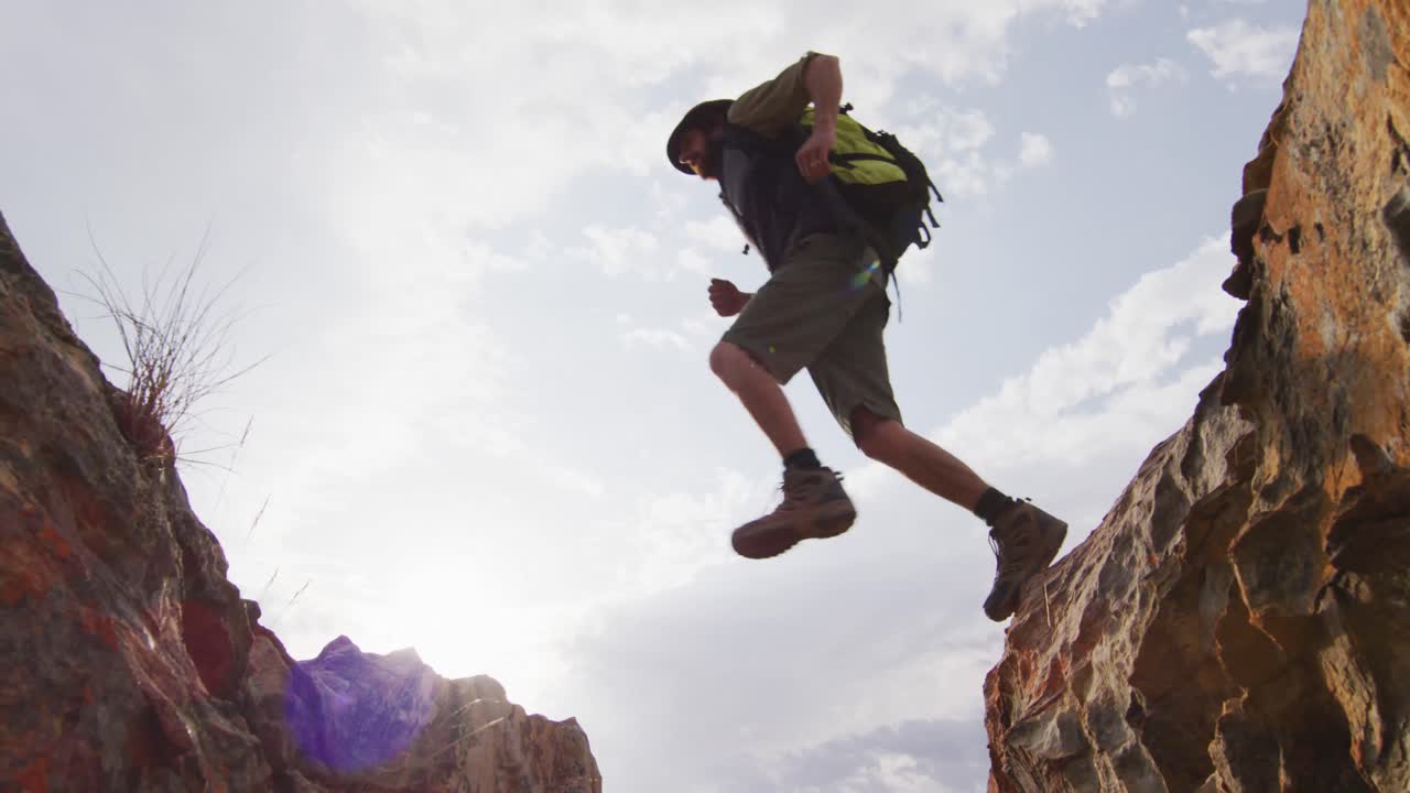 sobreviviente caucásico barbudo con mochila saltando a través de un barranco de montaña rocosa en el desierto
