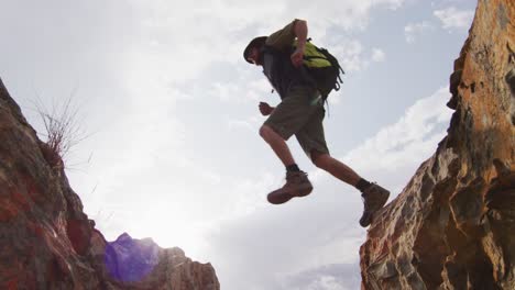Bearded-caucasian-male-survivalist-with-backpack-jumping-across-rocky-mountain-ravine-in-wilderness