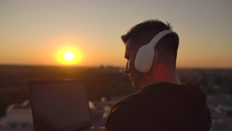 Hipster-man-with-a-laptop-on-the-edge-of-the-roof.-Freelancer-at-work.-Wireless-mobile-Internet.-He-works-on-the-Internet-with-headphones-and-listening-music