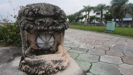 Hombre-Blanco-Obeso-Pasa-Junto-A-La-Estatua-Del-Perro-Shisa-En-Un-Parque-Público