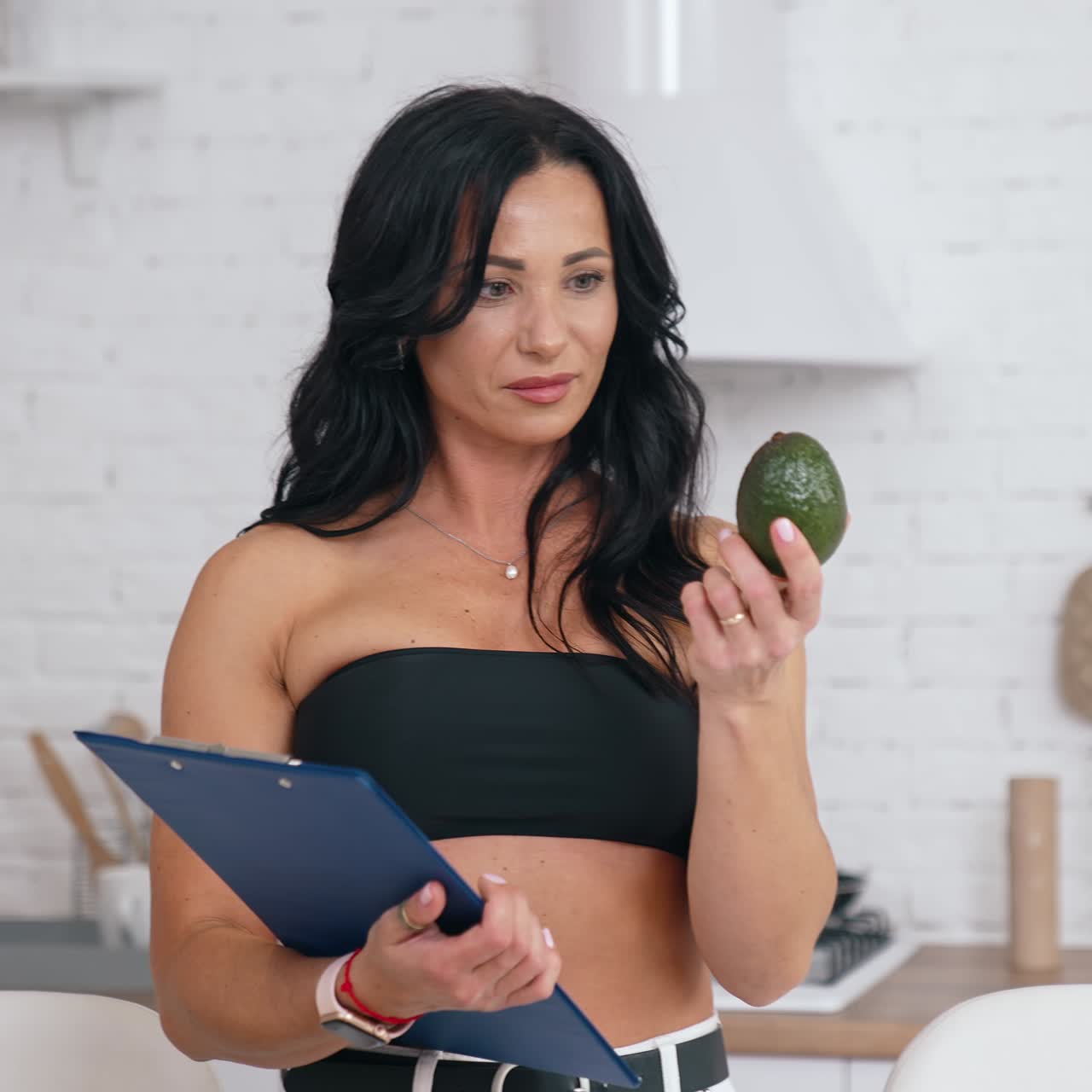 Pretty woman holding avocado at home. Beautiful housewife in short black top with a fresh fruit smiles on modern kitchen background