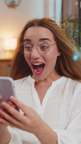 surprised young woman with windswept hair using smartphone winning lottery sitting on sofa at home