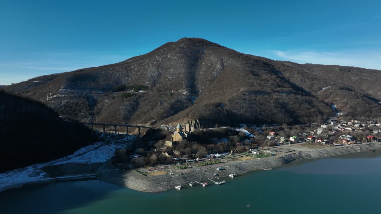a drone view of a village nestled at the base of a towering mountain, bordered by a calm lake. The serene setting highlights the harmony between the natural landscape and human habitation