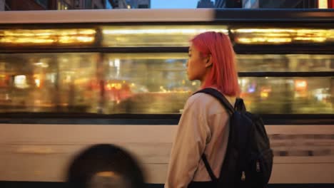 a young woman with pink hair waits by the street while a city bus speeds past her. the busy urban environment showcases vibrant lights and motion, capturing the essence of city life.