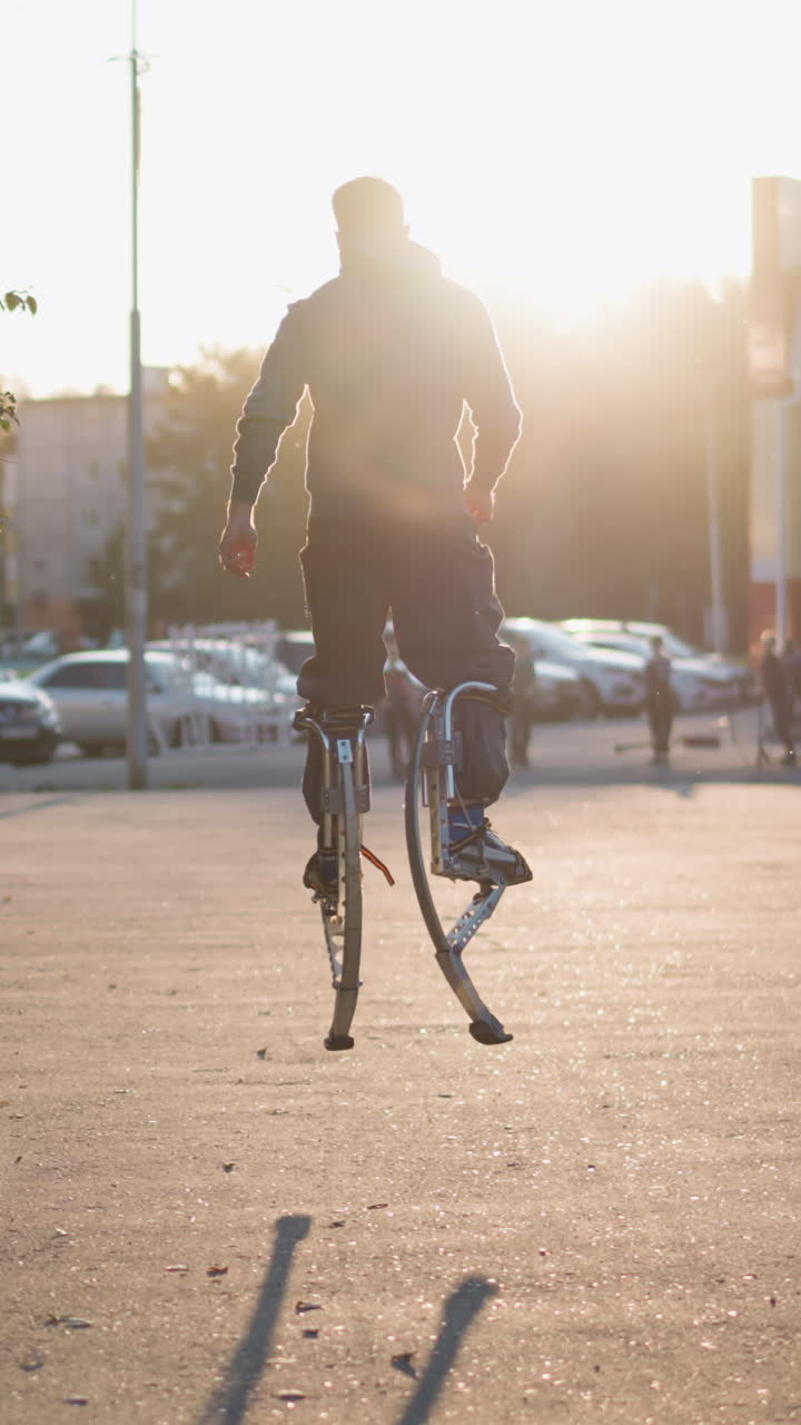 집, 나무, 사람들에 둘러싸인 주거 지역에서 해가 지는 동안 회색 후디와 어두운 회색 바지를 입고 기둥 위에 높이 뛰어다니는 chap의 뒷면