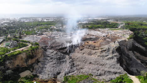 Aerial-view-around-a-junkyard-fire,-toxic-smoke-rising-in-middle-of-piles-of-junk,-in-Evento,-Santo-Domingo,-Dominican-Republic---Orbit,-drone-shot