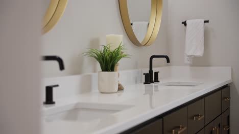 tight panning shot of a white bathroom countertop with 2 black faucets and a green plant