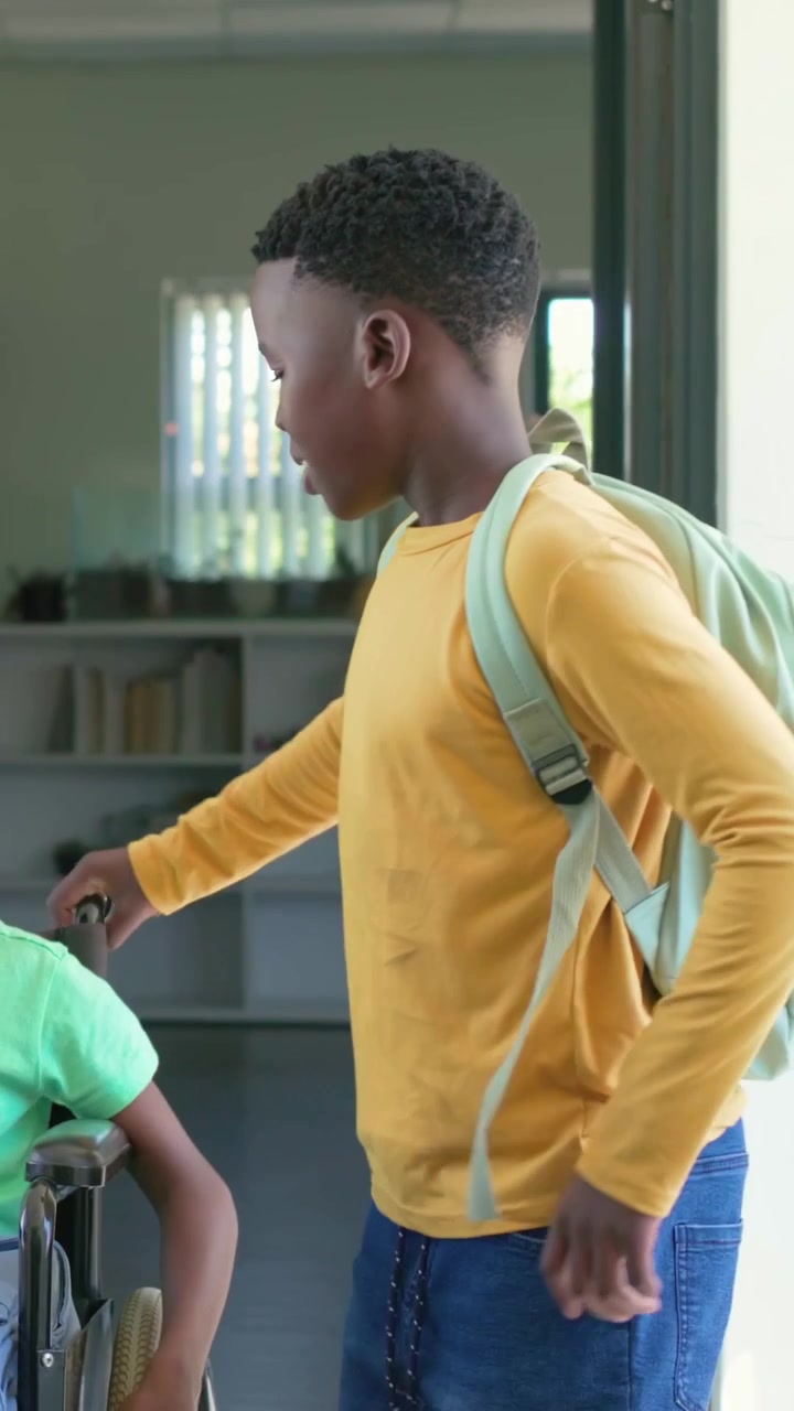 Vertical video: Coming door with backpack, yellow kid guiding wheelchair into classroom, copy space