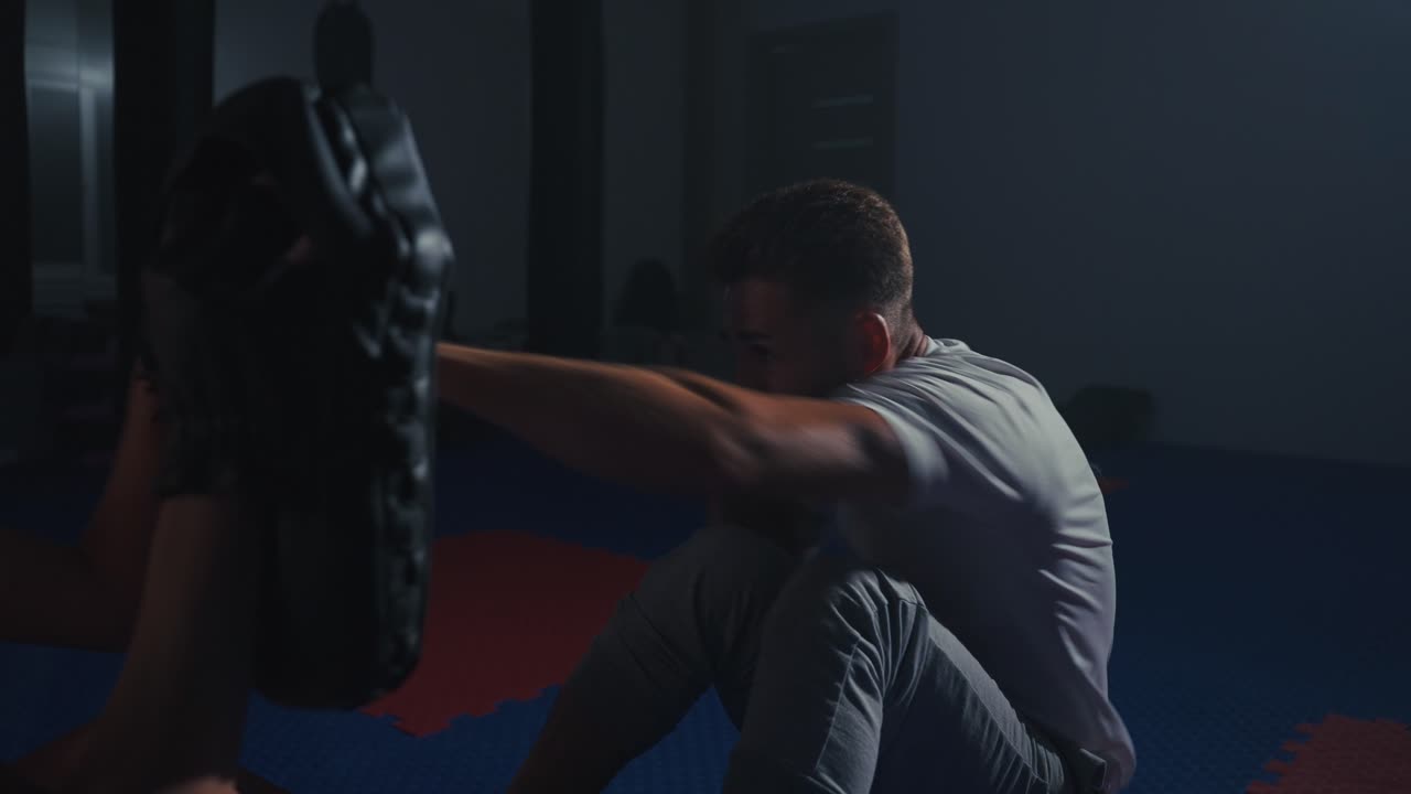 Man Doing Sit-Ups and Punching Pad with Trainer in Dark Gym