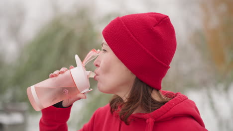 mom on maternity drinking water outdoors wearing red hoodie and beanie, holding pink bottle in serene natural setting with blurred background of greenery and light snow