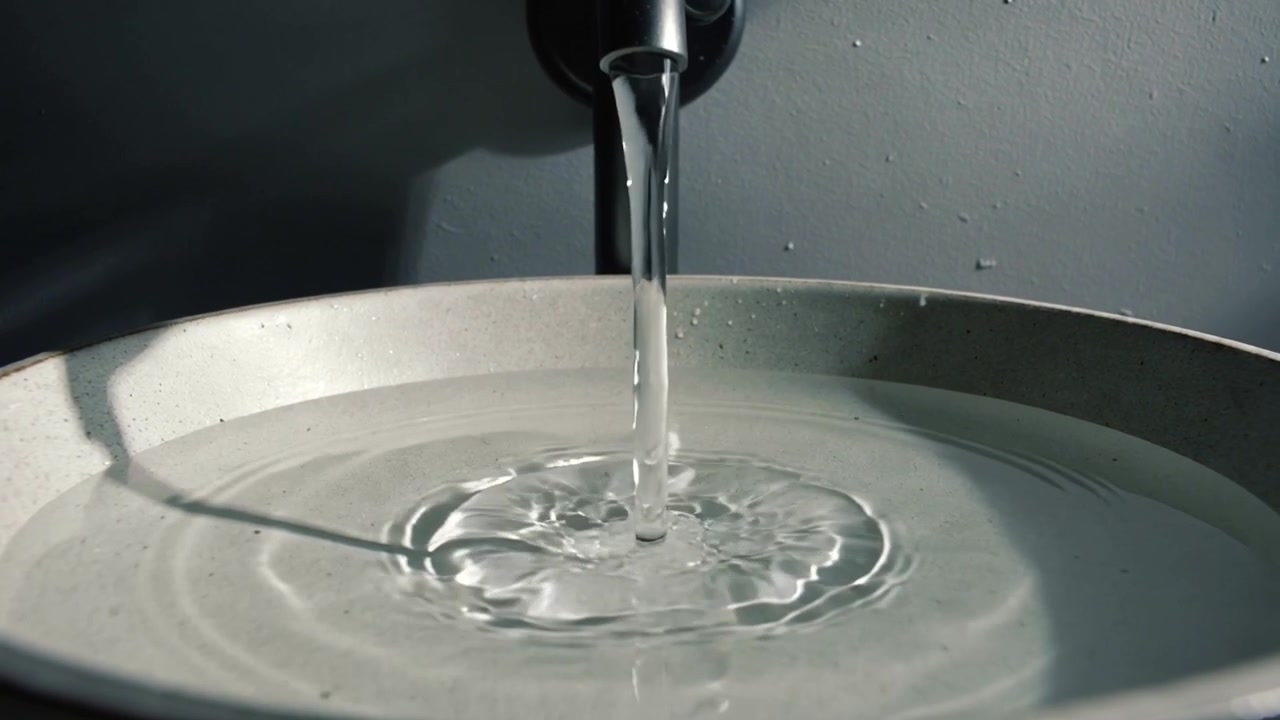 Apertura della valvola del rubinetto, beccuccio metallico a parete che versa un getto nel lavabo del bagno, formando increspature
