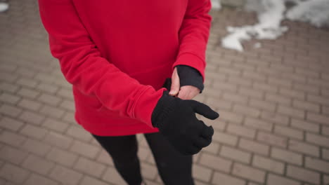 partial view of woman in red hoodie standing on interlocked path with snow along edges, putting on black winter gloves for warmth during cold weather in outdoor urban setting