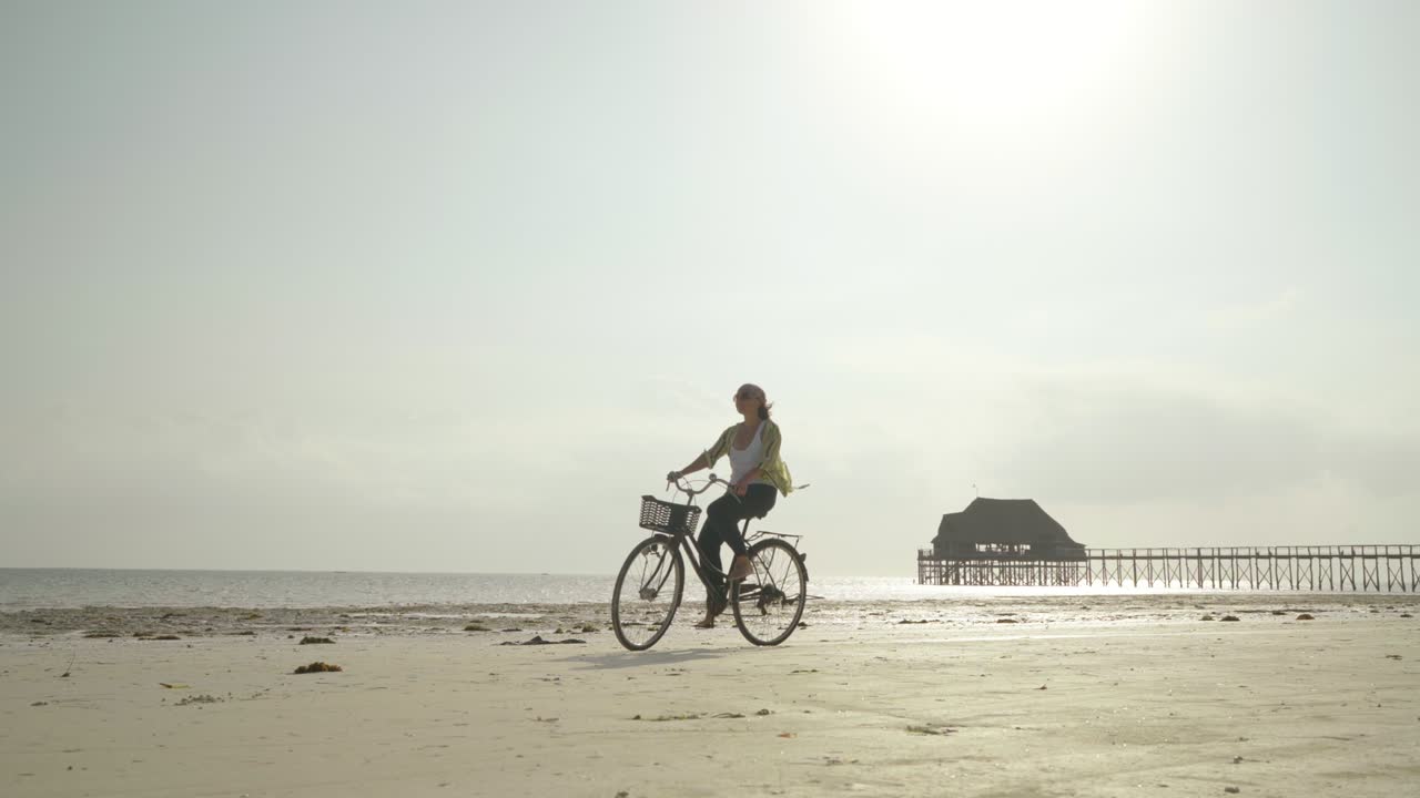 4K slow motion video of a Woman riding bicycle on sandy beach on sunny clear day near wooden pier. Biking concept, traveling, resort vacation concept.