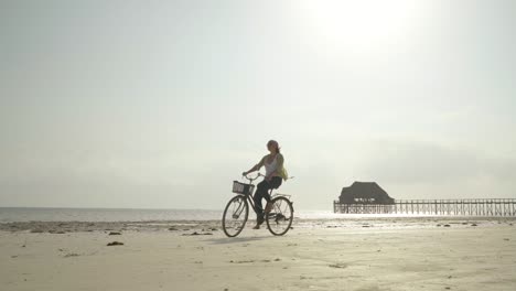 4k slow motion video of a woman riding bicycle on sandy beach on sunny clear day near wooden pier. biking concept, traveling, resort vacation concept.