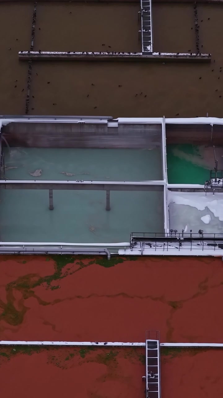 Vertical top-down drone footage of an industrial water treatment facility during winter, showing snow-covered infrastructure, geometric layouts, and essential urban water systems from above.