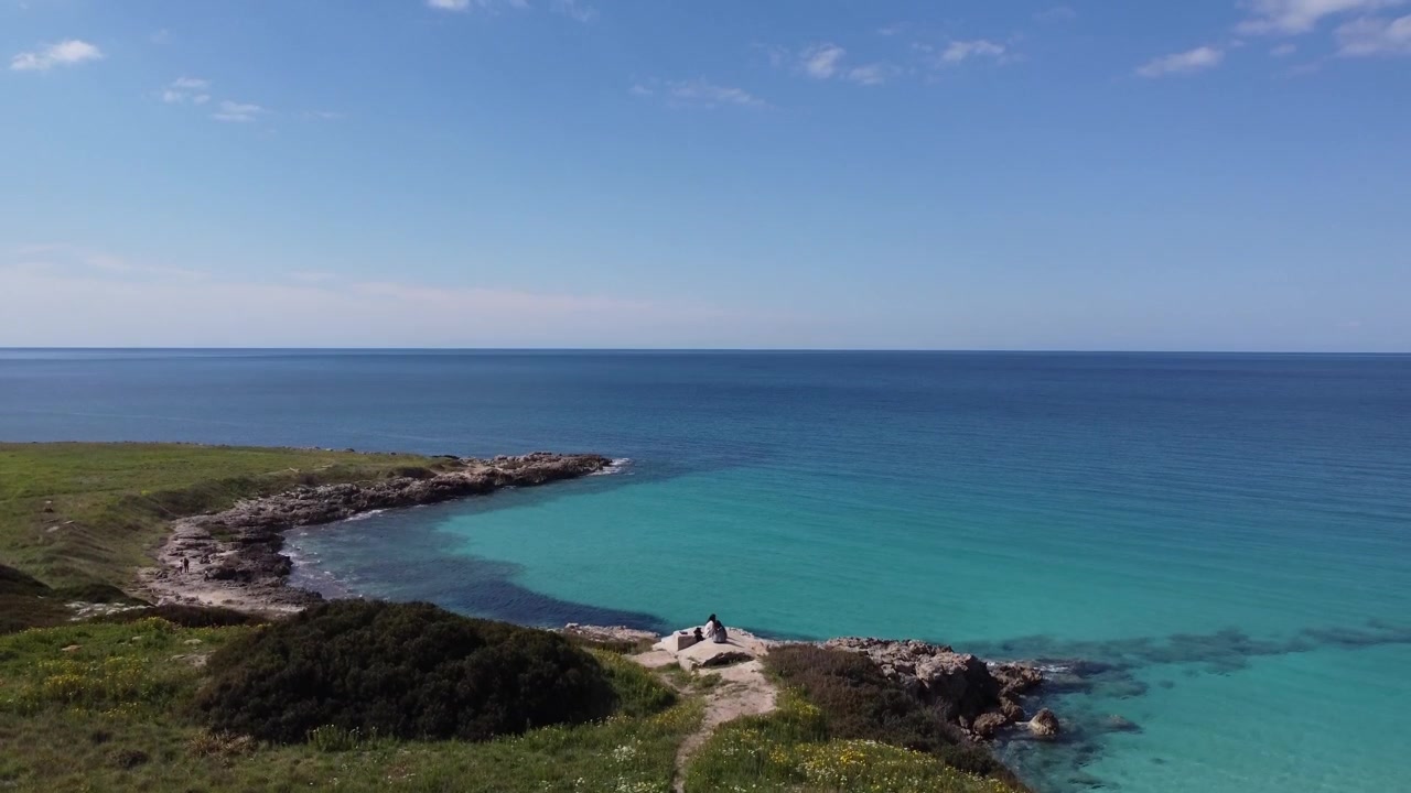 Ripresa aerea con drone di una costa rocciosa e frastagliata con acqua mediterranea limpida e turchese e una piccola penisola sotto un cielo blu, paesaggio costiero panoramico a Torre Sant'Andrea, Puglia, Italia.