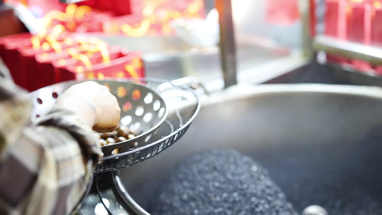 Vendor stirs roasted chestnuts in perforated pan over hot wok, vibrant market background, daylight
