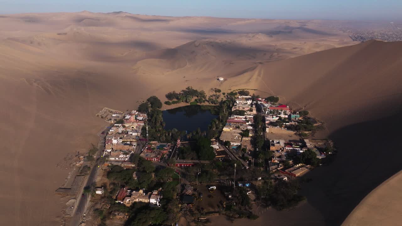 Aerial dolly shows oasis nestled beside steep dunes with soft light casting gentle shadows, establishing Huacachina resort town