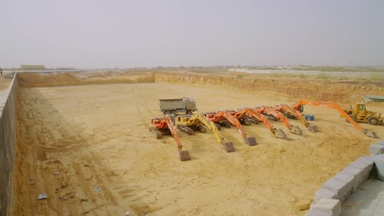 Wide shot of multiple excavators and a dump truck stationed at a deep rectangular construction site in Karachi, surrounded by sandy land and concrete bricks.