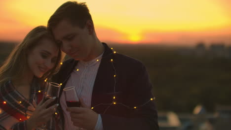 una joven pareja casada en el techo abrazándose y bebiendo vino rojo de vasos de pie vestidos de a cuadros y admirando la hermosa puesta de sol sobre la ciudad...