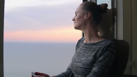 chica sonriente de pie junto a la ventana durante una puesta de sol y escuchando música con auriculares usando el teléfono móvil. disfrutando de la