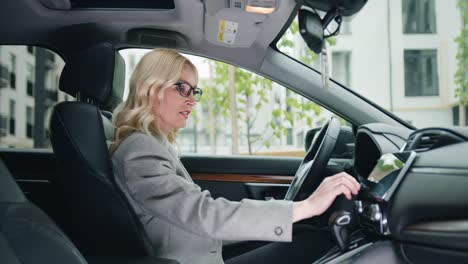 female mature driver in formal clothes adjusting radio dial button control, gps or air conditioning while sitting behind the whel of car. inside car view