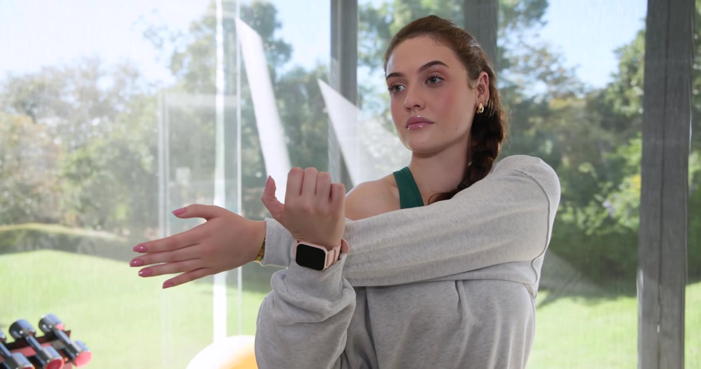 Donna adulta che si sistema la felpa, tirando un braccio sul petto, facendo stretching vicino alla finestra di una palestra casalinga e riscaldandosi.