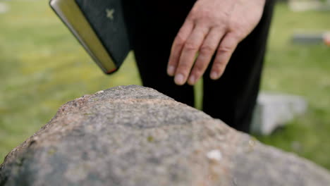 Nahaufnahme-Der-Hand-Eines-Mannes,-Der-Einen-Grabstein-Berührt,-Während-Er-Eine-Bibel-Auf-Einem-Friedhof-Hält-1