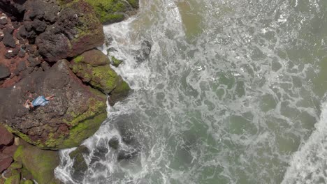 4k aerial top down drone shot of a beautiful 27-year-old young indian woman in white and blue dress relaxing on top of a cliff being crushed by the sea waves