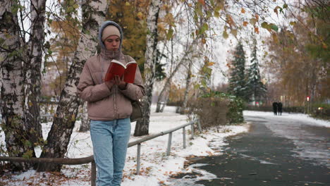 light skin lady flipping page of red book while standing quietly beside snowy path lined with birch trees and fallen leaves, dressed warmly in beige jacket and jeans during peaceful autumn afternoon
