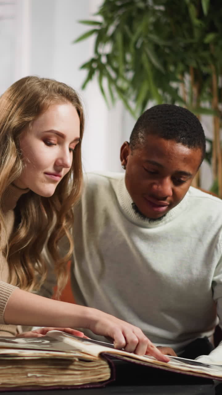 Cheerful lady shows family members on photos on large book to young African-American boyfriend on sofa in cozy living room slow motion