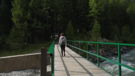 female hiker crosses sunny mountain river on wooden bridge